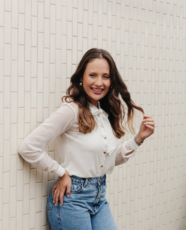 Victória de Souza sorrindo, apoiada em uma parede clara.